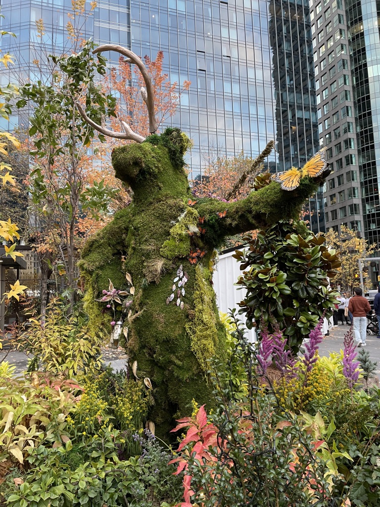 Upright topiary bear in Philadelphia, surrounded by flowers, butterfly in hand, wearing antlers.